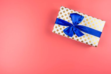 A gift wrapped in white paper with gold circles wrapped in a blue ribbon tied in a bow, isolated on a pink background, top view.