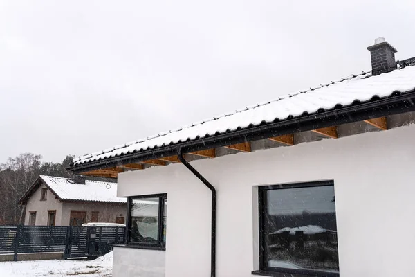 Single-family house roof covered with snow against a cloudy sky. Visible system chimney, roof trusses, windows and falling snow.