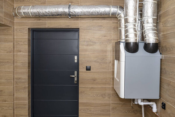 Home mechanical ventilation with heat recovery hanging on the wall in a modern gas boiler room with brown ceramic tiles imitating wood.