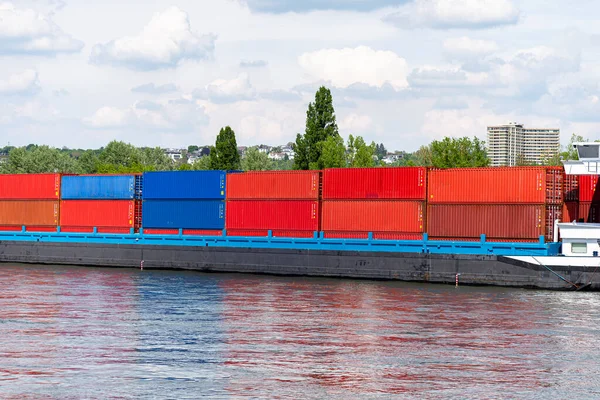 A barge carrying many containers on the Rhine in western Germany, trees and buildings in the background.