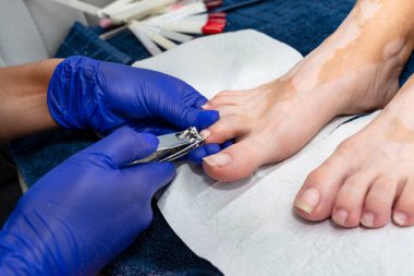 The manicurist cuts her toenails with a nail clipper wearing blue latex gloves.