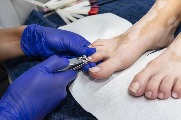 The manicurist cuts her toenails with a nail clipper wearing blue latex gloves.