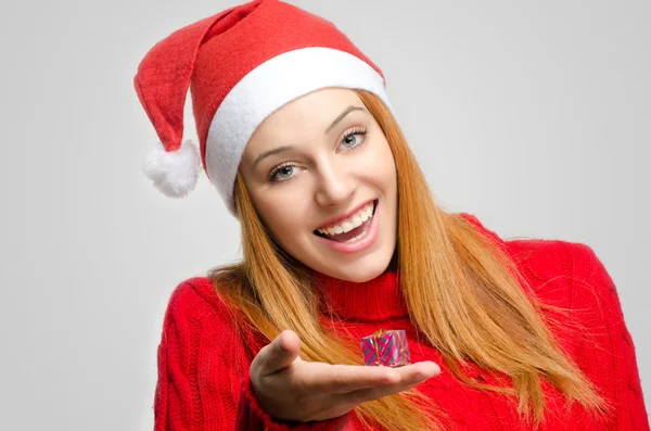 Crisis Christmas. Beautiful red hair woman holding a small Christmas present. Girl in red with Santa hat looking down smiling — Stock Photo, Image