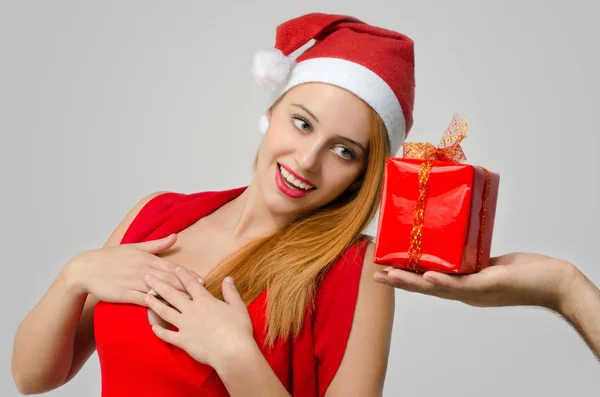 Hermosa mujer pelirroja recibiendo un regalo de Navidad. Chica en rojo con el sombrero de Santa sonriendo, la mano del hombre dando un regalo . — Foto de Stock