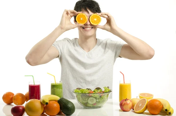 Gelukkig man houden voor zijn ogen sinaasappelen. vrolijke jonge man eten gezonde salade, fruit, jus d'orange en groene smoothie. — Stockfoto