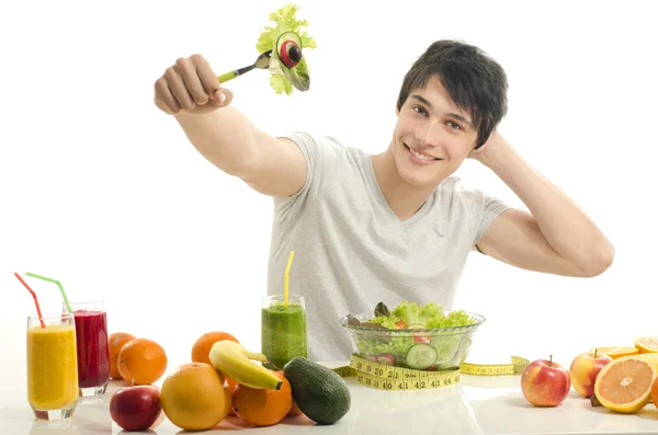 Счастливчик за столом, полным органических продуктов, соков и смузи. Веселый молодой человек ест здоровый салат и фрукты. Изолированные на белом . — стоковое фото