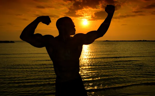 Silueta de culturista posando al amanecer en la playa, entrenando por la mañana, hombre fuerte mostrando sus músculos — Foto de Stock