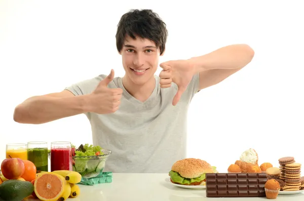 Hombre eligiendo entre frutas, batidos y alimentos orgánicos saludables contra dulces, azúcar, un montón de dulces y una gran hamburguesa, alimentos poco saludables —  Fotos de Stock