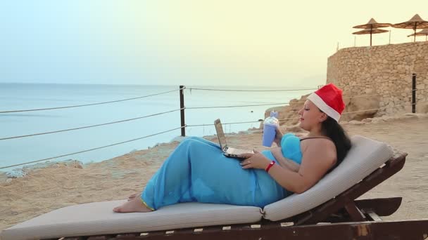 Μια γυναίκα στην παραλία σε μια ξαπλώστρα με ένα μαγιό παρεό και ένα καπέλο του Άη Βασίλη πίνει ένα κοκτέιλ και συγχαίρει τους φίλους για το νέο έτος από ένα φορητό υπολογιστή — Αρχείο Βίντεο