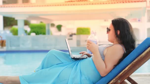 Une femme en maillot de bain et un paréo sur une chaise longue près de la piscine boit un cocktail et travaille à distance — Video