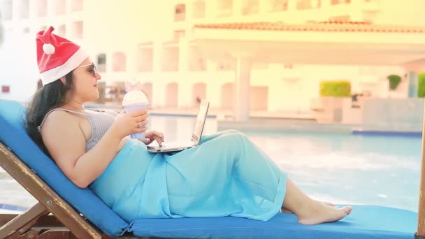 圣诞假期期间，一位戴着圣爪帽躺在游泳池边晒太阳的女人通过视频通话，喝着鸡尾酒 — 图库视频影像
