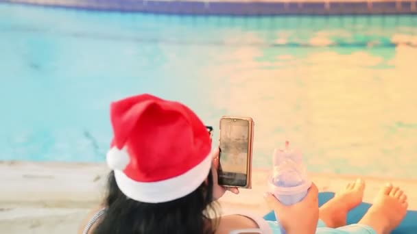 Een vrouw in een ligstoel bij het zwembad in een kerstmuts tijdens de kerstvakantie communiceert in een smartphone en drinkt een cocktail. Glans van zonlicht. schieten van achteren — Stockvideo