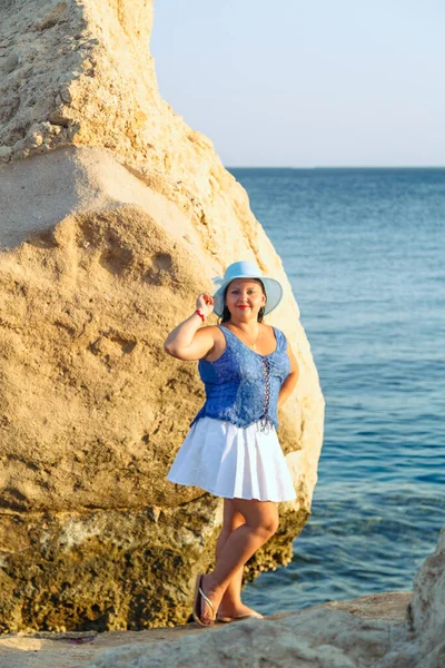 Una joven judía con una falda blanca y un sombrero azul en la orilla del mar cerca de las rocas. — Foto de Stock