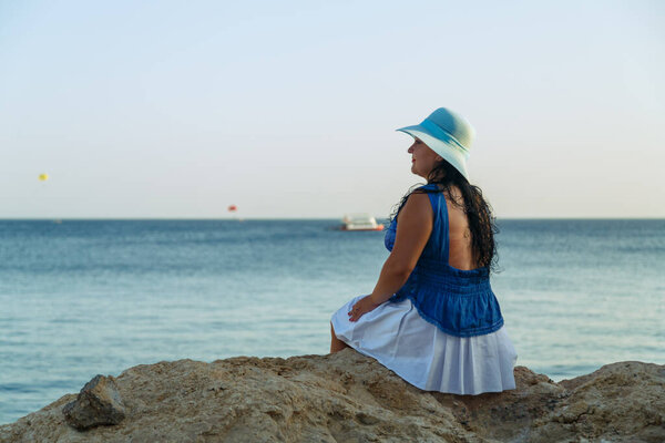 A young brunette woman in a white skirt and blue hat on the seashore sits with her back to the camera. Horizontal photo