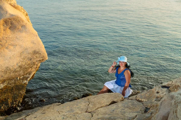 Μια νεαρή γυναίκα με λευκή φούστα και ένα καπέλο του ήλιου στην παραλία κάθεται σε ένα βράχο θαυμάζοντας τη φύση. — Φωτογραφία Αρχείου
