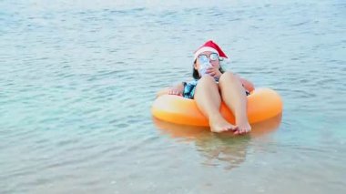Noel Baba şapkalı ve güneş gözlüklü esmer bir kadın Noel tatilinde denizde bir çember içinde yüzer ve uzaklığa bakar..