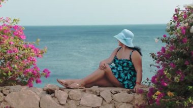 Çiçeklerle sarılmış taştan bir duvarda oturan ve denize hayran esmer bir kadın.