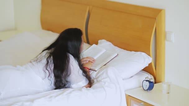 ベッドの中で白い衣を着たブルネットの女性は彼女の胃の上に横たわっている間本を読む — ストック動画