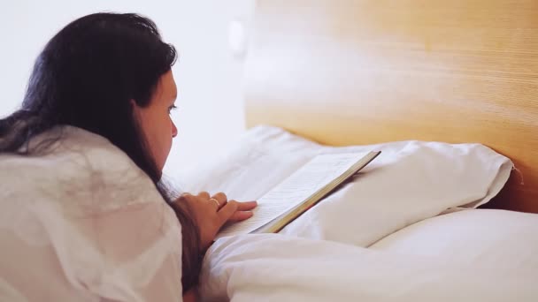 Μια γυναίκα με άσπρη ρόμπα σε μεγάλο κρεβάτι διαβάζει ένα βιβλίο πυροβολώντας από πίσω.. — Αρχείο Βίντεο