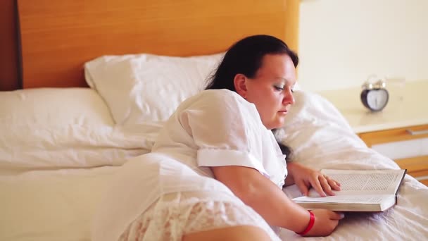 Żydówka w białym szlafroku w łóżku czyta książkę leżąc na boku — Wideo stockowe