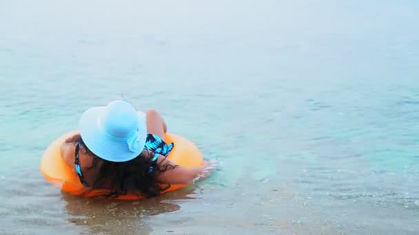 Una morena con sombrero azul y gafas de sol nada en un círculo en el mar cerca de la orilla y bebe un cóctel. — Vídeos de Stock