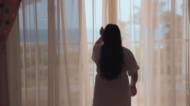 En brunette kvinde i en hvid kåbe om morgenen åbner gardinerne og går ud på balkonen med udsigt over havet. Optagelse fra bagsiden – Stock-video