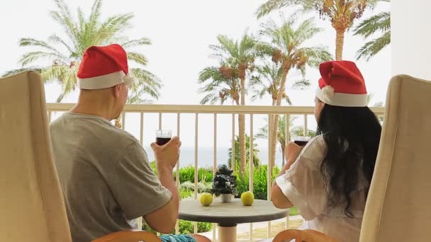 Een getrouwd stel in de kerstman hoeden viert Kerstmis op het balkon met uitzicht op de zee, drink een cocktail op de tafel, een kleine kerstboom — Stockvideo