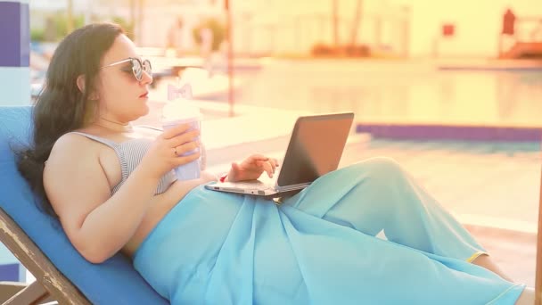 Une femme en maillot de bain et lunettes de soleil sur une chaise longue près de la piscine travaille à distance avec un ordinateur portable — Video