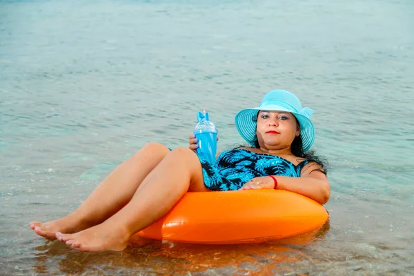 Een vrouw met een strohoed en een bril in de zee in een zwemcirkel met een cocktail in haar hand. — Stockfoto