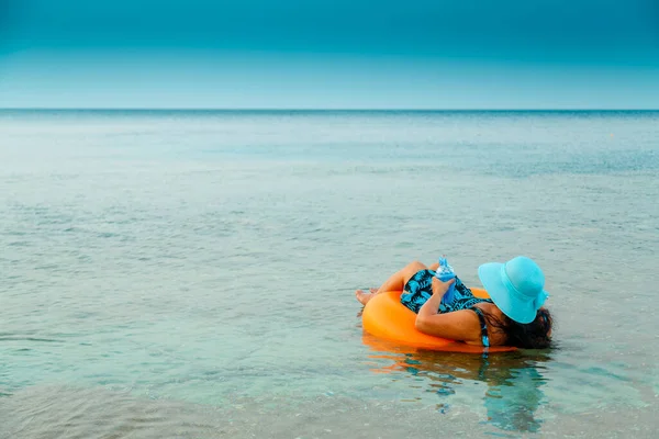 Μια γυναίκα με ψάθινο καπέλο και γυαλιά στη θάλασσα σε έναν κύκλο κολύμβησης με ένα κοκτέιλ στη θέα του χεριού της από πίσω. — Φωτογραφία Αρχείου