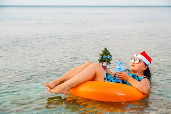 Женщина в плавательном кольце в шляпе Санта Клауса с коктейлем и маленькой елкой в руке плавает в море во время рождественских каникул. — стоковое фото