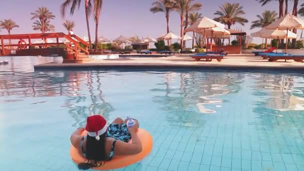 Havuzda yüzme çemberinde Noel Baba şapkalı bir kadın, arka planda palmiye ağaçları ve güneşli yerlerde kameraya arkası dönük bir şekilde kokteyl içiyor. — Stok video