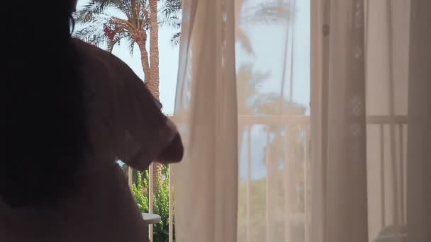 En brunett kvinna på morgonen öppnar gardinerna och går ut till balkongen med utsikt över havet. Skjuta bakifrån — Stockvideo