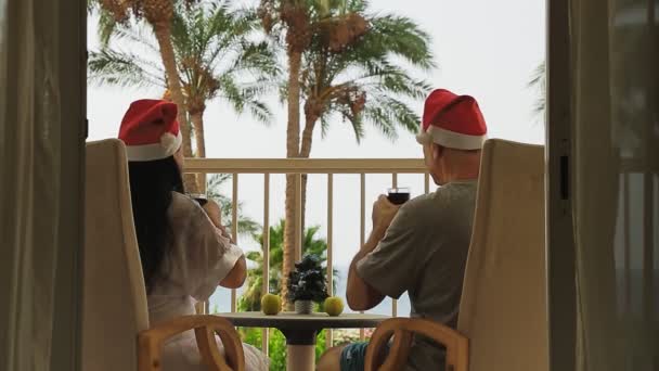 Een getrouwd stel in de kerstman hoeden vieren Kerstmis en Nieuwjaar op het balkon met uitzicht op zee op de tafel een kleine kerstboom — Stockvideo