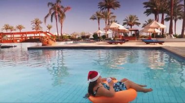 Havuzda yüzme çemberinde Noel Baba şapkalı bir kadın, palmiye ağaçlarının ve dinlenme yerlerinin arka planında tatilde kokteyl içiyor. Genel plan