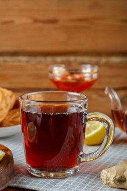 Masanın üzerinde bir fincan koyu çay, arka planda limon ve zencefil, çörekler ve vazoda reçel var. Dikey fotoğraf