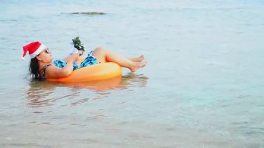 Noel Baba şapkalı ve güneş gözlüklü esmer bir kadın Noel tatilinde denizde daireler çizerek yüzer ve kokteyl içer. Genel plan