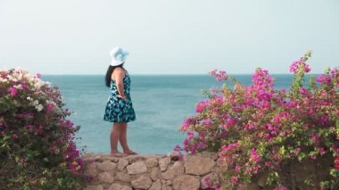 Güneşlik elbisesi ve şapkası olan esmer bir kadın rüzgarda çiçeklerle çevrili taş bir duvarda duruyor ve denize bakıyor. Genel plan