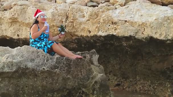 サンタクラスの帽子とサングラスのブルネットの女性が岩の上に座って海を見て クリスマス休暇中にカクテルを飲みます 計画全体 — ストック動画