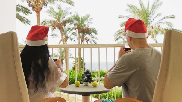 一对戴着圣诞老人帽子的已婚夫妇在阳台上俯瞰大海庆祝圣诞节 他们在桌上喝着鸡尾酒 一棵小小的圣诞树 总体计划 — 图库视频影像
