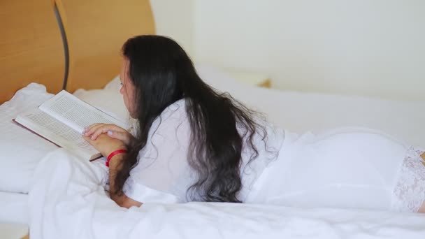 Брюнетка в білому халаті в ліжку читає книгу, лежачи на животі. Бічний постріл . — стокове відео