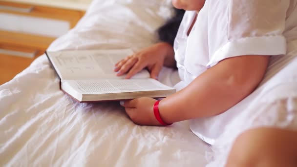 Una donna con una vestaglia bianca sul letto sta leggendo un libro appoggiato ai gomiti per un'iniezione senza volto — Video Stock