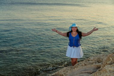 Beyaz etekli ve güneş şapkalı genç bir kadın gün batımında kollarını açmış denizin arka planına karşı ayakta duruyor..