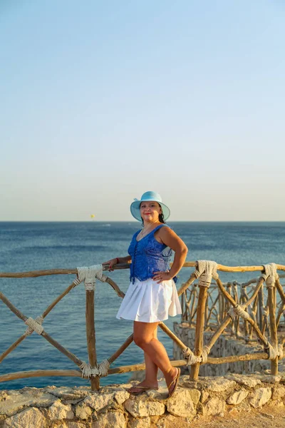 Mujer morena joven en una falda blanca y sombrero azul en la orilla del mar feliz. — Foto de Stock
