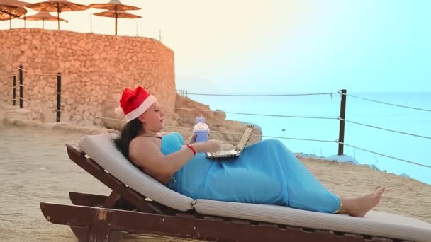 Una donna bruna in riva al mare in un lettino in un cappello di Babbo Natale beve un cocktail e stampa su un computer portatile — Video Stock