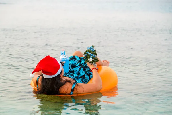 A woman in a swimming ring in a Santa Claus hat with a cocktail and a small Christmas tree in her hand floats in the sea. Shooting from the back. Horizontal photo
