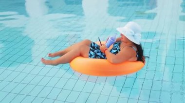 Yüzme havuzunda güneş şapkalı ve mayo giymiş bir kadın kokteyl içer ve dinlenir. Genel plan