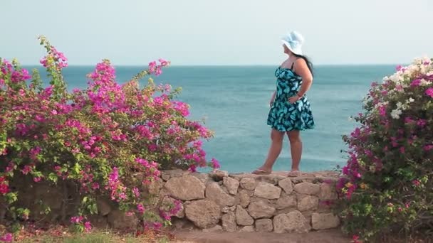 Una mujer judía en un sarafán y un sombrero están parados en una pared de piedra entrelazada con flores y mira al mar. — Vídeos de Stock