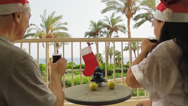 Ženatý pár v Santa Claus klobouky slaví Vánoce na balkóně u moře a pít koktejl. — Stock video