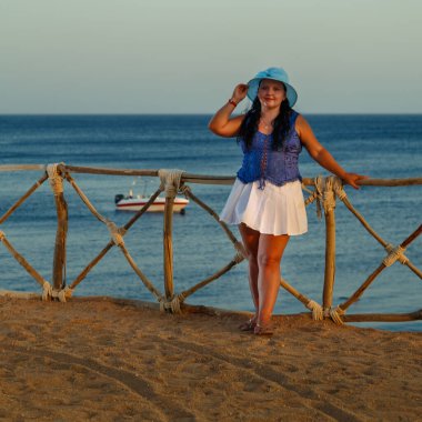 Beyaz etekli ve güneş şapkalı genç bir kadın gün batımında denizin arka planında kıyıda duruyor..